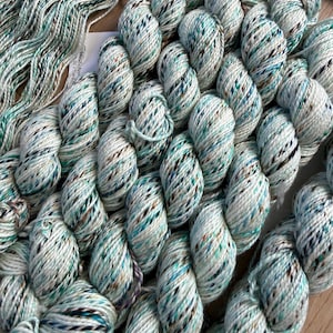 Puede incluir: Múltiples madejas de hilo con una paleta de colores variados en blanco, verde azulado claro y motas de color marrón oscuro. El hilo está retorcido en paquetes compactos, mostrando una textura de fibra multicolor con motas visibles.