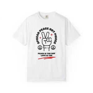 Puede incluir: Camiseta blanca con un gráfico negro y rojo. El gráfico presenta una mano haciendo el signo de la paz, rodeada de las palabras "SPREAD PEACE NOT PIECES" y "PEACE IS THE WAY LOVE IS THE".
