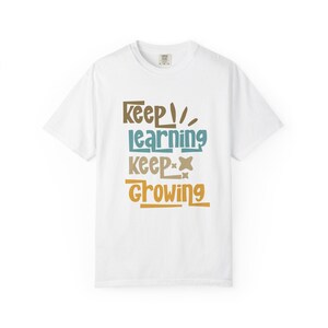 Puede incluir: Camiseta blanca con la frase "Keep Learning Keep Growing" en letras marrones, turquesas y doradas. El texto está apilado. La camiseta tiene cuello redondo y mangas cortas. Ropa informal.