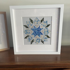 May include: A framed artwork featuring a blue and white floral design in a starburst pattern. The artwork is framed in a white frame and is displayed on a wooden surface.