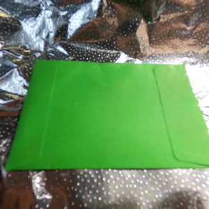 May include: A bright green envelope, likely for cards or letters, is centered in the image. The envelope is rectangular with a flap on the right side. The background is a shiny, silver material with a dotted pattern.