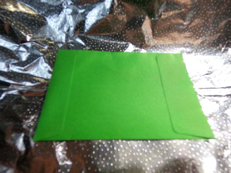 May include: A bright green envelope, likely for cards or letters, is centered in the image. The envelope is rectangular with a flap on the right side. The background is a shiny, silver material with a dotted pattern.