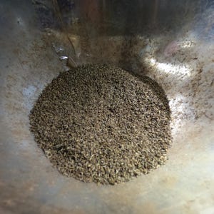 May include: Close-up of a pile of small, dark brown seeds inside a silver metal bowl. The seeds are densely packed, creating a textured surface. The bowl has a slightly curved shape.