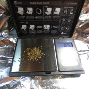 May include: A digital pocket scale with a black frame and a silver weighing surface. The scale displays a weight of 0.26, with a small pile of dried plant matter on the surface. The top of the scale has a user guide.