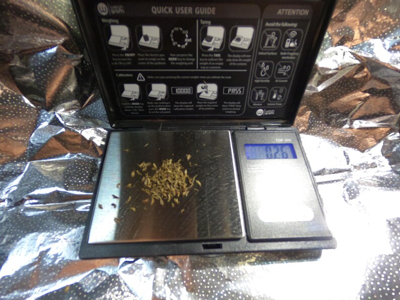May include: A digital pocket scale with a black frame and a silver weighing surface. The scale displays a weight of 0.26, with a small pile of dried plant matter on the surface. The top of the scale has a user guide.