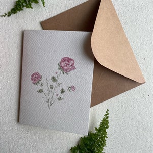 May include: A blank white card with a watercolor illustration of pink roses and green leaves. The card is sitting on a white surface with a brown envelope and green ferns.