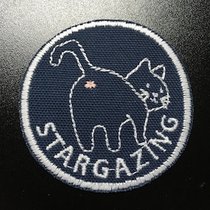 May include: Embroidered patch with a navy blue background and a white stitched cat design. The cat is facing away, with a small pink detail. The words "STARGAZING" are stitched in white around the edge of the patch.