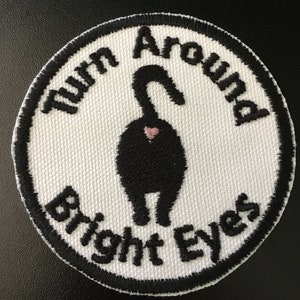 May include: A round, white patch with black embroidered text and a black cat rear end illustration. The text reads "Turn Around Bright Eyes" in a circular pattern. A small pink heart is visible.