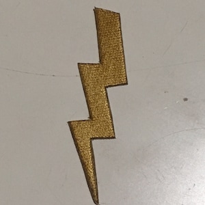 May include: A gold-colored lightning bolt patch with a textured surface. The patch is a stylized representation of a lightning bolt, with sharp angles and a pointed bottom. The patch is likely for decorative purposes.