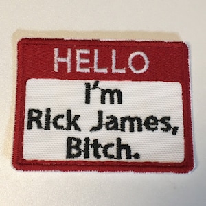 May include: A rectangular patch with a red border and a white center. The text "HELLO" is at the top, with "I'm Rick James, Bitch." below in black letters. The patch is likely for clothing or accessories.