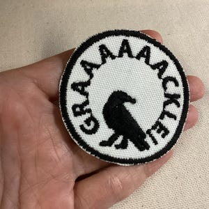 May include: A circular embroidered patch featuring a white background with a black border. In the center, a black bird silhouette is depicted. The text GRAAAAACKLE is embroidered in black capital letters along the upper curved edge of the patch.
