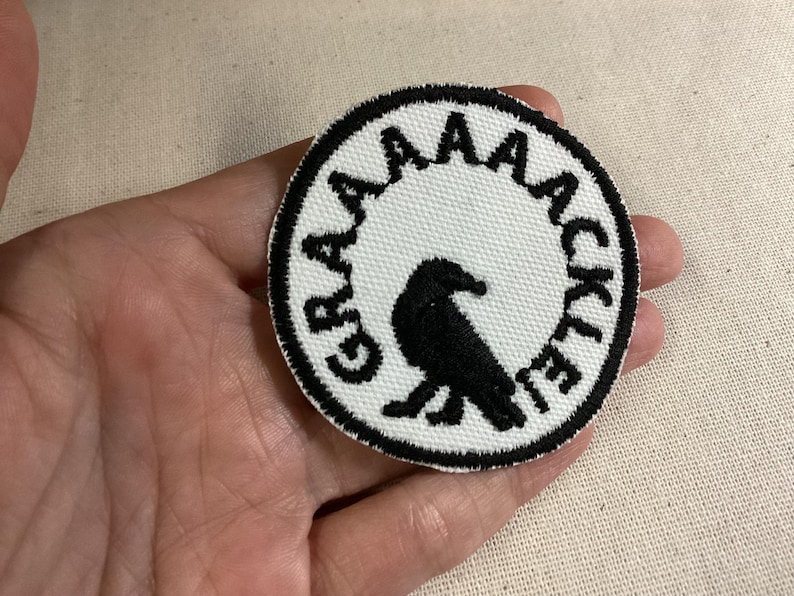 May include: A circular embroidered patch featuring a white background with a black border. In the center, a black bird silhouette is depicted. The text GRAAAAACKLE is embroidered in black capital letters along the upper curved edge of the patch.
