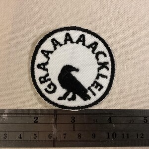 May include: A circular embroidered patch featuring a black bird silhouette in the center on a white background. The text GRAAAAACKLE is stitched in black along the inner border. A metal ruler is positioned below, showing a diameter of 2 inches.