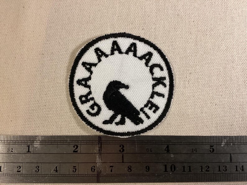 May include: A circular embroidered patch featuring a black bird silhouette in the center on a white background. The text GRAAAAACKLE is stitched in black along the inner border. A metal ruler is positioned below, showing a diameter of 2 inches.
