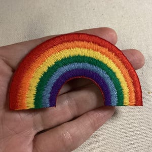 May include: A semi-circular embroidered patch featuring a vibrant rainbow design. The patch displays concentric bands of red, orange, yellow, green, blue, and purple thread, creating a classic arc shape with a clean, stitched border finish.