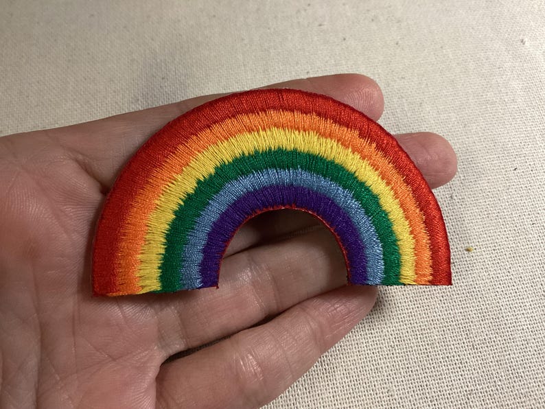 May include: A semi-circular embroidered patch featuring a vibrant rainbow design. The patch displays concentric bands of red, orange, yellow, green, blue, and purple thread, creating a classic arc shape with a clean, stitched border finish.