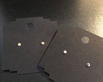 May include: Two pairs of small, circular, metallic stud earrings are displayed on individual black rectangular cardboard display cards. Each card features a circular punch-out hole at the top center for hanging, with the earrings positioned below.