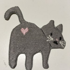May include: A gray cat-shaped patch with a pink heart on its back. The cat has white whiskers, black eyes, and a pink nose. The patch is made of a textured fabric and is likely an iron-on or sew-on applique.