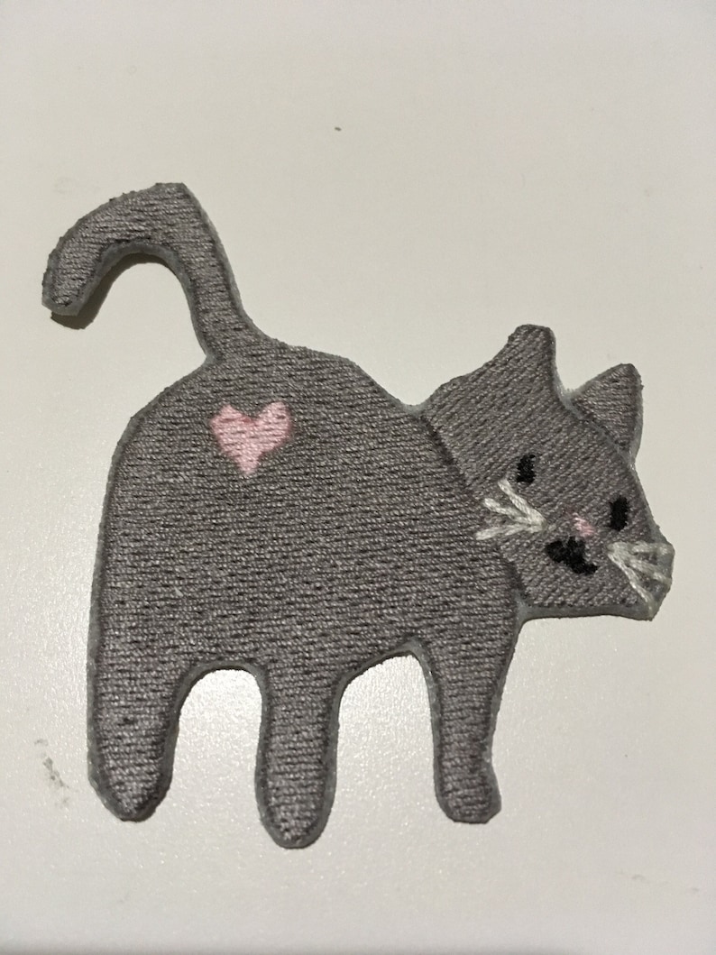 May include: A gray cat-shaped patch with a pink heart on its back. The cat has white whiskers, black eyes, and a pink nose. The patch is made of a textured fabric and is likely an iron-on or sew-on applique.