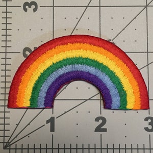 May include: A semi-circular rainbow embroidered patch featuring concentric bands of red, orange, yellow, green, light blue, and purple thread. The patch is positioned over a cutting mat with a visible ruler showing a width of 2.5 inches.
