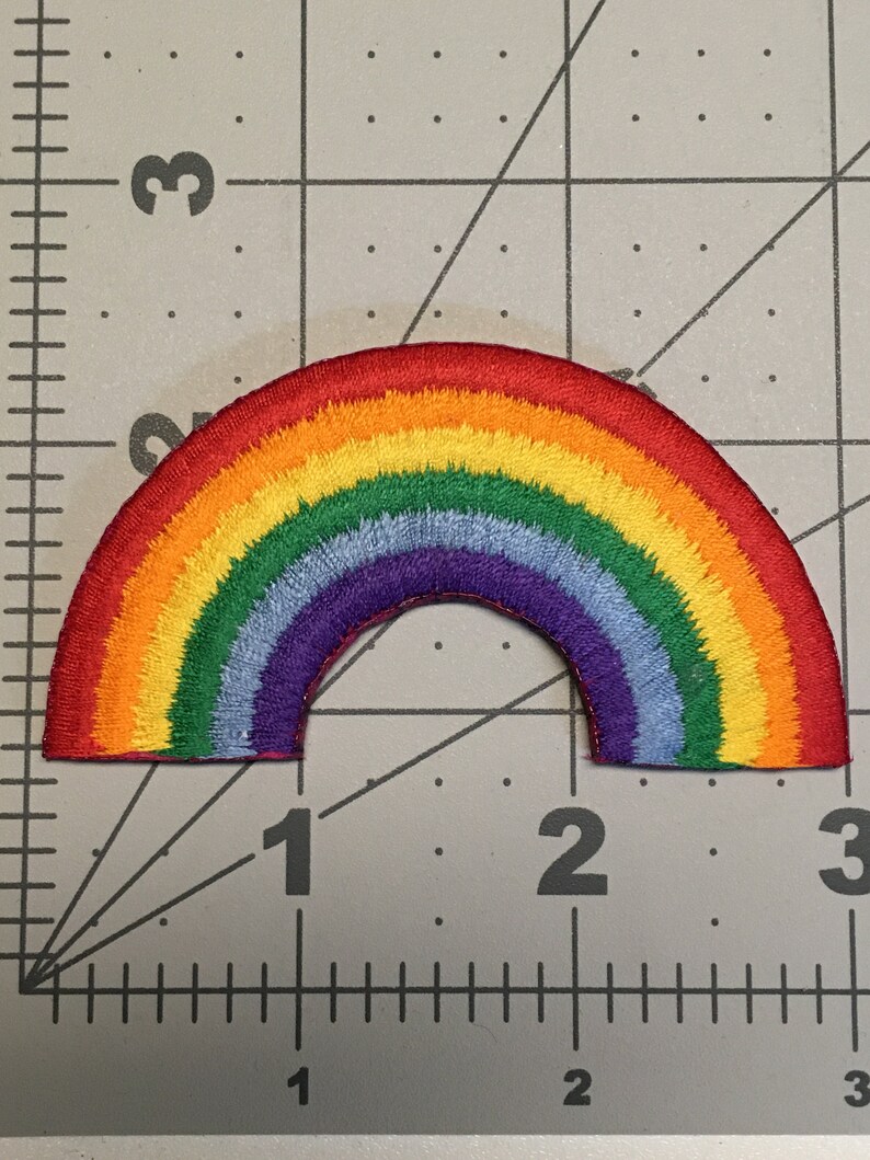 May include: A semi-circular rainbow embroidered patch featuring concentric bands of red, orange, yellow, green, light blue, and purple thread. The patch is positioned over a cutting mat with a visible ruler showing a width of 2.5 inches.