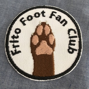 May include: A circular embroidered patch with a white background and a black border. The center features a brown animal paw with light pink pads. The text "Frito Foot Fan Club" is embroidered in black lettering along the upper curved edge.