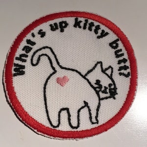 May include: A round, white patch with a red border features a black outline of a cat's rear end with a pink heart. The text "What's up kitty butt?" is in black, arched around the cat image. The patch is likely for clothing or accessories.