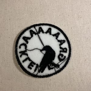 May include: A circular embroidered patch featuring a white background with a black border. The design includes a black silhouette of a bird perched on a line, surrounded by the black text "AAAAACK" arranged in a circular pattern around the edge.