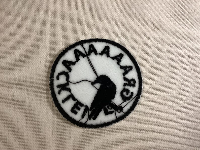 May include: A circular embroidered patch featuring a white background with a black border. The design includes a black silhouette of a bird perched on a line, surrounded by the black text "AAAAACK" arranged in a circular pattern around the edge.