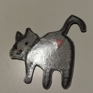 May include: A gray cat-shaped patch with a heart on its rear. The patch has a textured surface and is viewed from the back, showing the cat's tail and legs. The cat's face has pink cheeks and black eyes.
