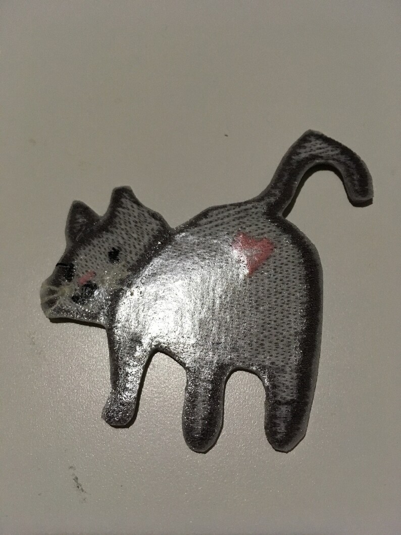 May include: A gray cat-shaped patch with a heart on its rear. The patch has a textured surface and is viewed from the back, showing the cat's tail and legs. The cat's face has pink cheeks and black eyes.