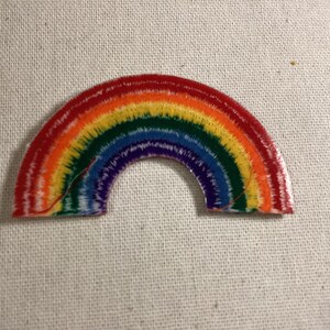 May include: A semi-circular embroidered patch featuring a vibrant rainbow design. The arc displays concentric bands of red, orange, yellow, green, blue, and purple threads with a textured, stitched appearance throughout the entire surface.