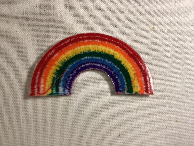 May include: A semi-circular embroidered patch featuring a vibrant rainbow design. The arc displays concentric bands of red, orange, yellow, green, blue, and purple threads with a textured, stitched appearance throughout the entire surface.