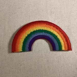 May include: A semi-circular embroidered patch featuring a vibrant rainbow design. The arc consists of concentric bands of red, orange, yellow, green, blue, and purple thread, creating a textured, colorful pattern on a fabric base.
