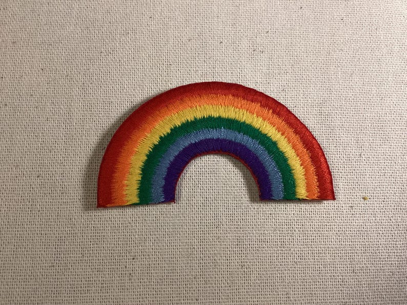 May include: A semi-circular embroidered patch featuring a vibrant rainbow design. The arc consists of concentric bands of red, orange, yellow, green, blue, and purple thread, creating a textured, colorful pattern on a fabric base.