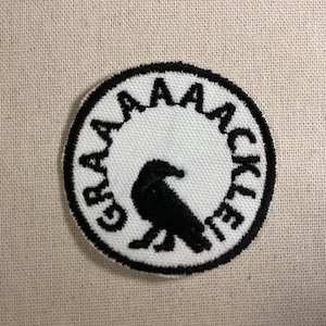 May include: A circular embroidered patch featuring a white background with a black border. In the center is a black silhouette of a bird. The text GRAAAAACKLE is embroidered in black capital letters along the inner edge of the circular border.
