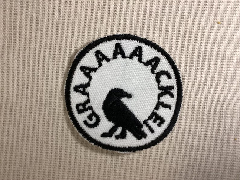 May include: A circular embroidered patch featuring a white background with a black border. In the center is a black silhouette of a bird. The text GRAAAAACKLE is embroidered in black capital letters along the inner edge of the circular border.