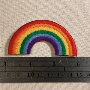 May include: A semi-circular rainbow embroidered patch featuring red, orange, yellow, green, blue, and purple thread. A metal ruler is positioned below the patch, showing it spans approximately 3.5 inches in width from end to end.