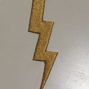 May include: Gold-colored lightning bolt patch. The patch has a textured surface and a zig-zag design. The lightning bolt is a bright gold color and is likely made of fabric or a similar material. The patch is a decorative item.