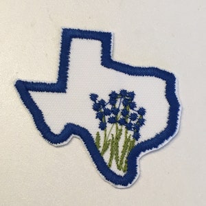 May include: Embroidered patch in the shape of Texas, featuring a white interior and a blue outline. The patch showcases bluebonnet flowers with green stems and leaves, a symbol of Texas.