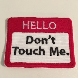 May include: A rectangular patch with a red border and a white center. The word "HELLO" is embroidered in white at the top, and below it, the phrase "Don't Touch Me" is embroidered in black.
