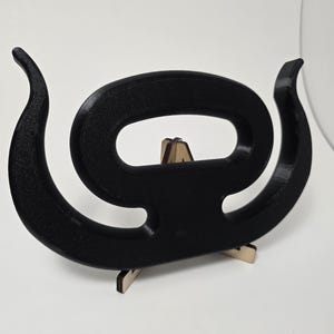 May include: A black, abstract sculpture in the shape of a stylized letter 'G' or a spiral. The sculpture is made of a textured material and is supported by a small, light-colored wooden stand. The design has curved, elongated ends.