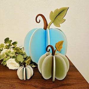 Op de afbeelding: Drie houten pompoenversieringen in verschillende maten. De grootste is lichtblauw, de middelste is groen en de kleinste is wit. Elk heeft een bruine stengel en een groen of geel blad. De pompoenen worden tentoongesteld op een donkerbruin oppervlak.