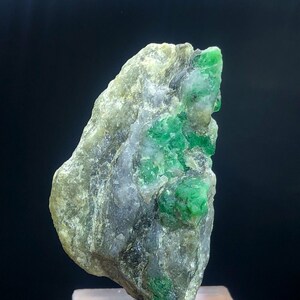May include: A rough, gray rock with a cluster of bright green emeralds embedded in it. The rock is on a clear display stand.