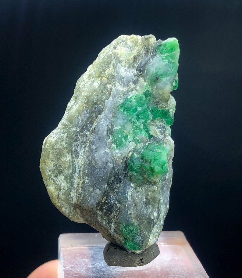 May include: A rough, gray rock with a cluster of bright green emeralds embedded in it. The rock is on a clear display stand.