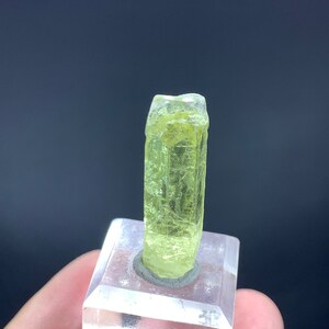 Puede incluir: Un esp&eacute;cimen de cristal amarillo p&aacute;lido montado sobre una base de acr&iacute;lico transparente. El cristal es largo y delgado con una punta puntiaguda.