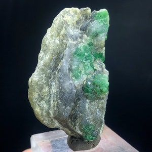 May include: A rough, gray and white rock with a cluster of bright green emeralds embedded in it. The rock is on a clear display stand.
