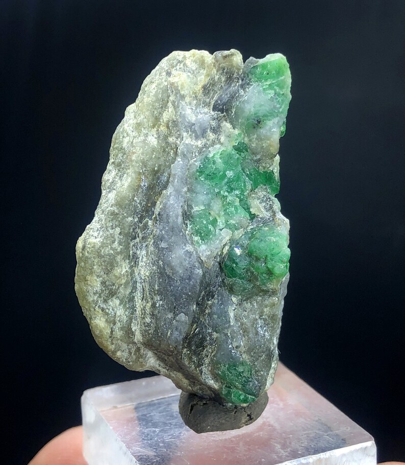 May include: A rough, gray and white rock with a cluster of bright green emeralds embedded in it. The rock is on a clear display stand.