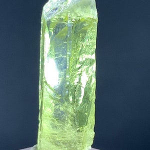 Puede incluir: Un cristal de color verde amarillo brillante con una forma alargada y facetada. El cristal est&aacute; sentado sobre una superficie gris.