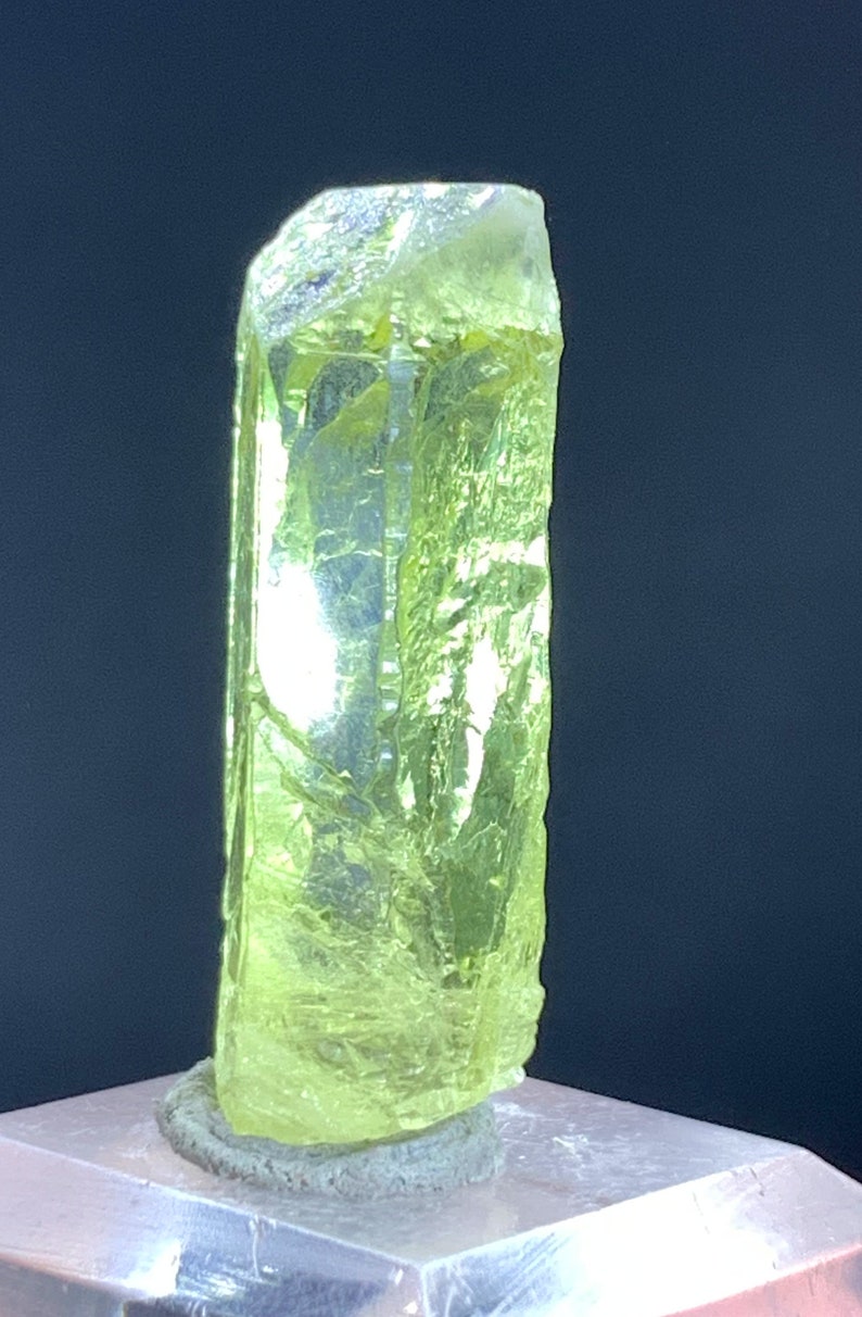 Puede incluir: Un cristal de color verde amarillo brillante con una forma alargada y facetada. El cristal est&aacute; sentado sobre una superficie gris.